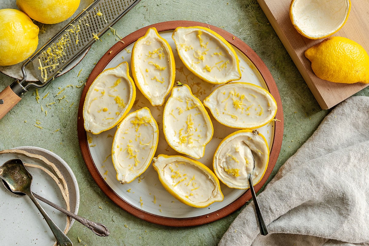 Halved lemon shells filled with lemon mousse are arranged on a plate, with lemon zest sprinkled on top. A grater, lemon, and spoon are nearby.