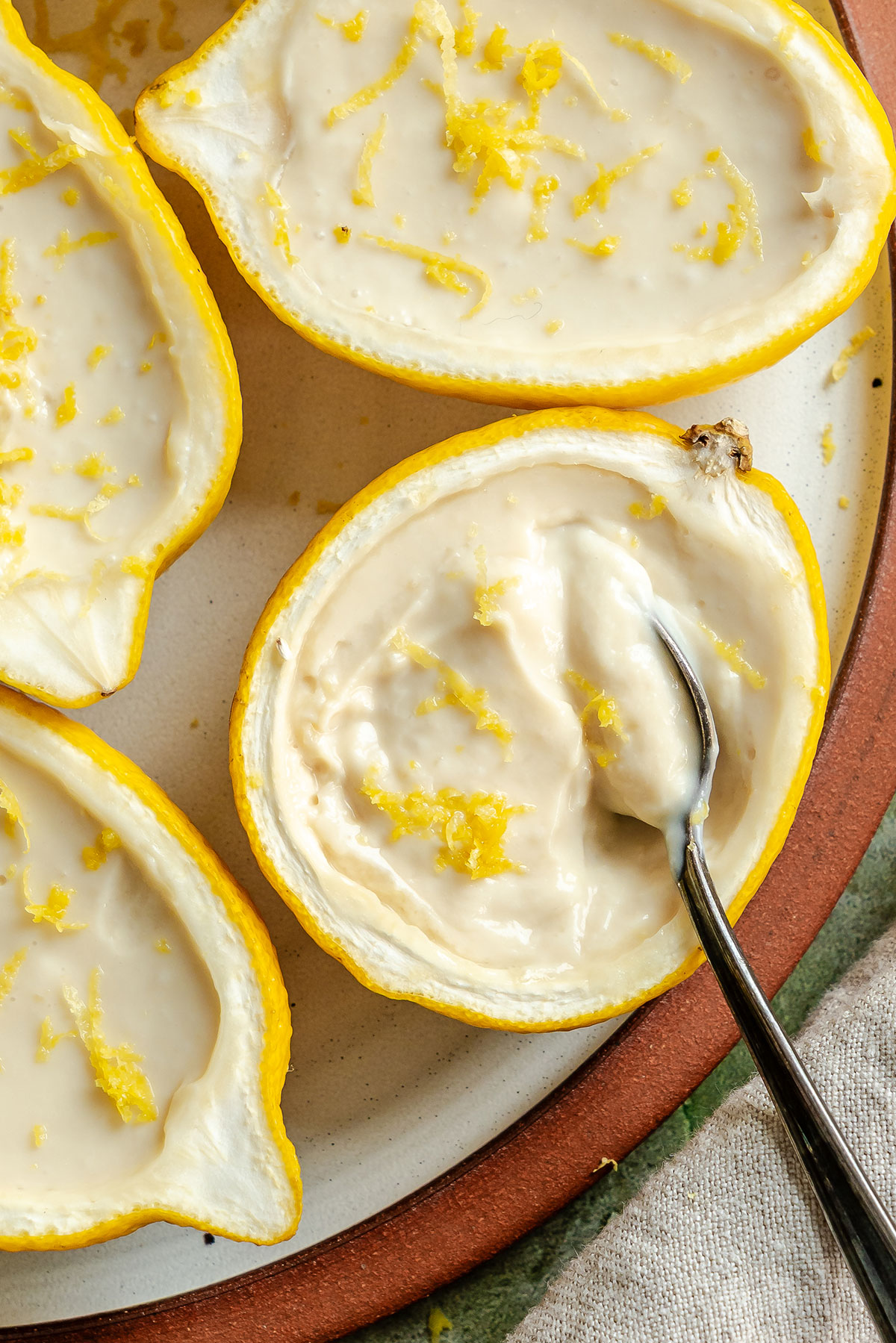 Halved lemons filled with creamy lemon dessert, garnished with lemon zest, with a spoon scooping from one half on a plate.