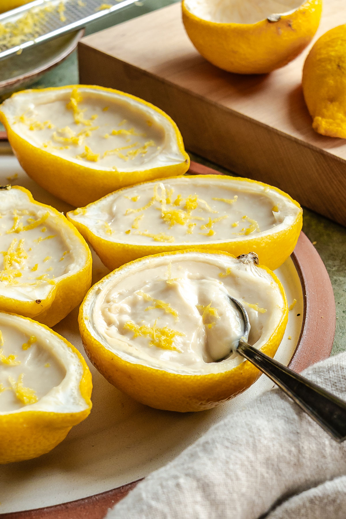 Halved lemons filled with creamy dessert, topped with lemon zest, arranged on a plate with a spoon in one half and a wooden cutting board in the background.