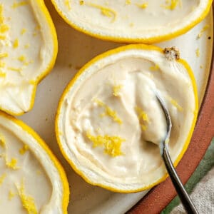 Close-up of creamy Vegan Lemon Posset served in hollowed-out lemon halves, garnished with lemon zest, with a spoon scooping some out—this vegan dessert gets its silky texture from silken tofu.