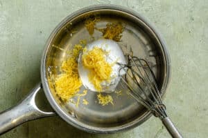 A metal saucepan containing whipped cream and lemon zest with a metal whisk on a green countertop.