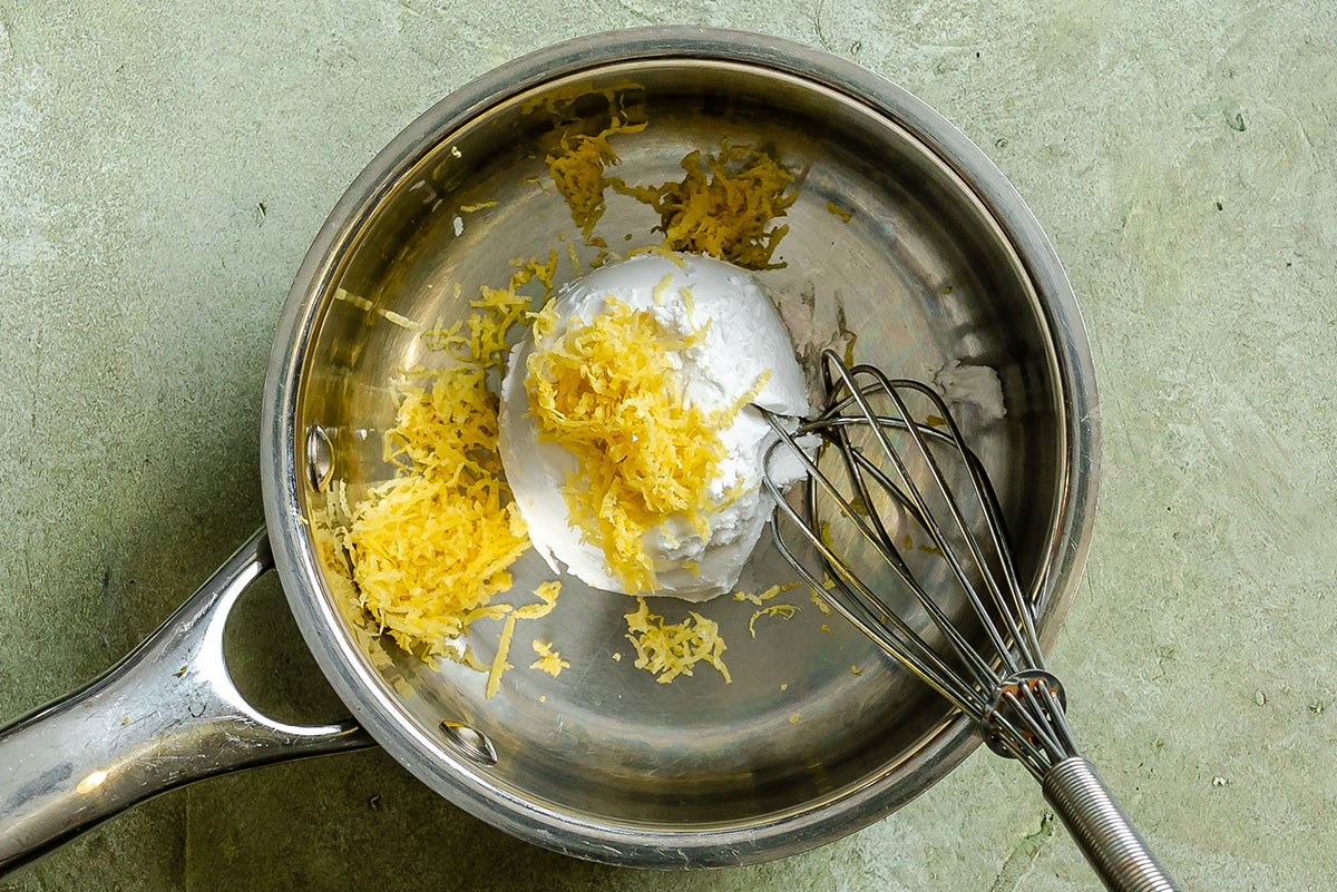 A metal saucepan containing whipped cream and lemon zest with a metal whisk on a green countertop.