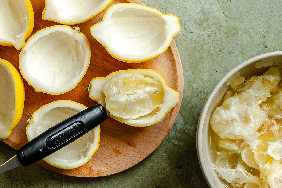 Lemon halves with pulp removed are on a wooden board; a bowl with lemon pulp and seeds sits nearby, and a spoon rests inside one lemon half.
