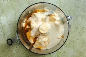 A food processor bowl contains white and beige ingredients including sugar, yogurt, and honey, ready to be blended.
