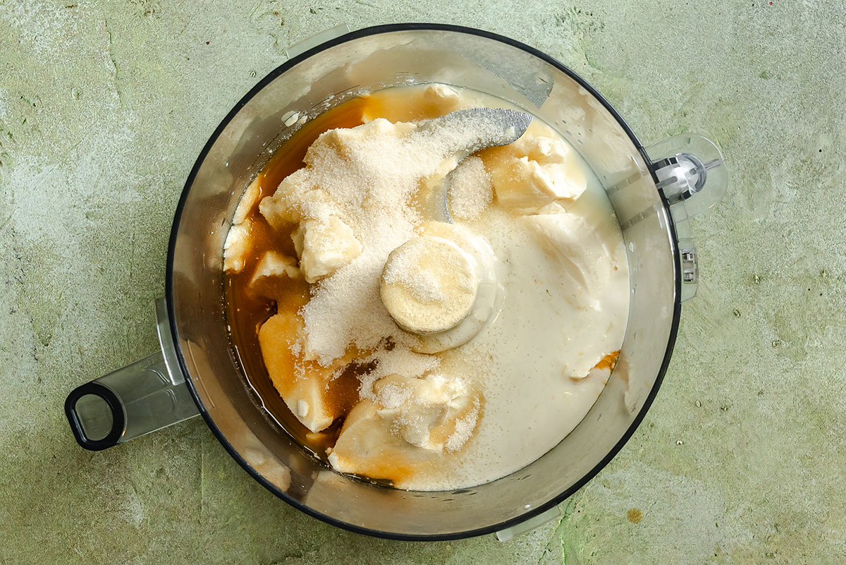 A food processor bowl contains white and beige ingredients including sugar, yogurt, and honey, ready to be blended.