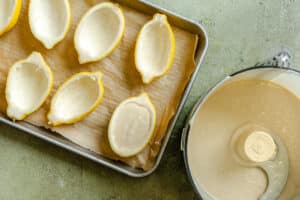 Halved and hollowed lemon shells on a tray next to a food processor bowl filled with creamy mixture, ready to be filled.