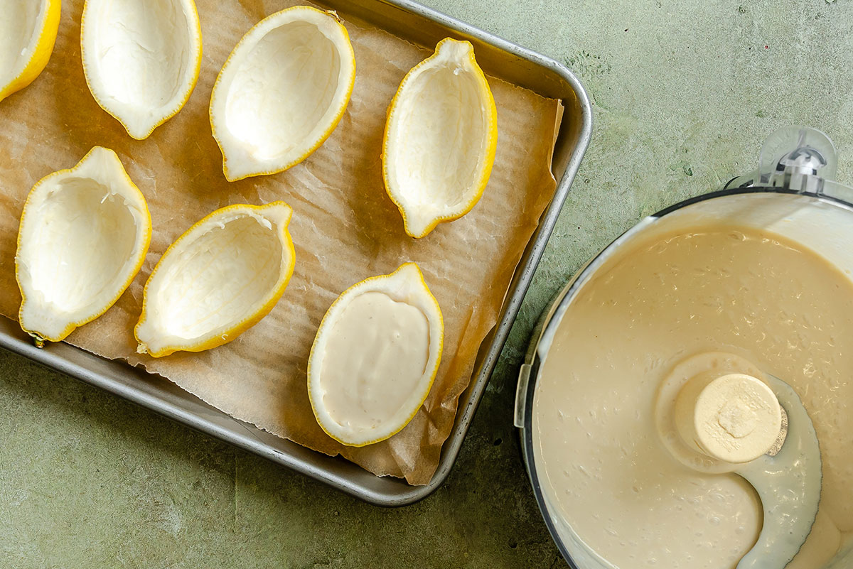 Halved and hollowed lemon shells on a tray next to a food processor bowl filled with creamy mixture, ready to be filled.