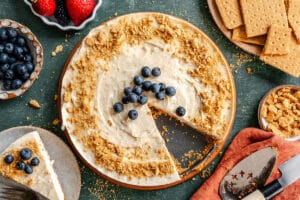 Overhead view of a cheesecake topped with blueberries and crumbled graham crackers, with a slice removed and placed on a plate nearby. Surrounding are bowls of fruit and extra graham crackers.