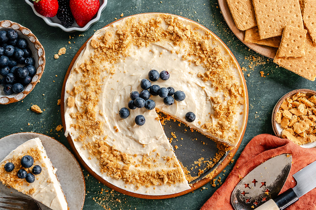Overhead view of a cheesecake topped with blueberries and crumbled graham crackers, with a slice removed and placed on a plate nearby. Surrounding are bowls of fruit and extra graham crackers.