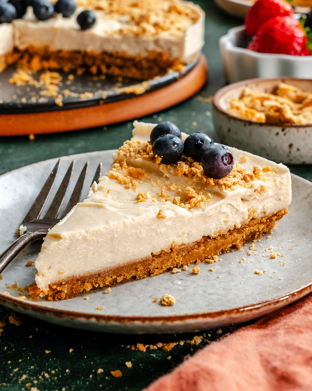 A slice of cheesecake with a graham cracker crust, topped with blueberries and crumbs, on a plate with a fork. The remaining cheesecake and a bowl of berries are in the background.