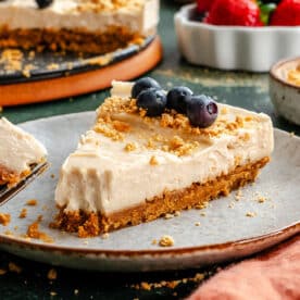 A slice of cheesecake with a crumbly crust, topped with blueberries and crumbs, sits on a plate with a fork. A whole cheesecake and a bowl of mixed berries are in the background.
