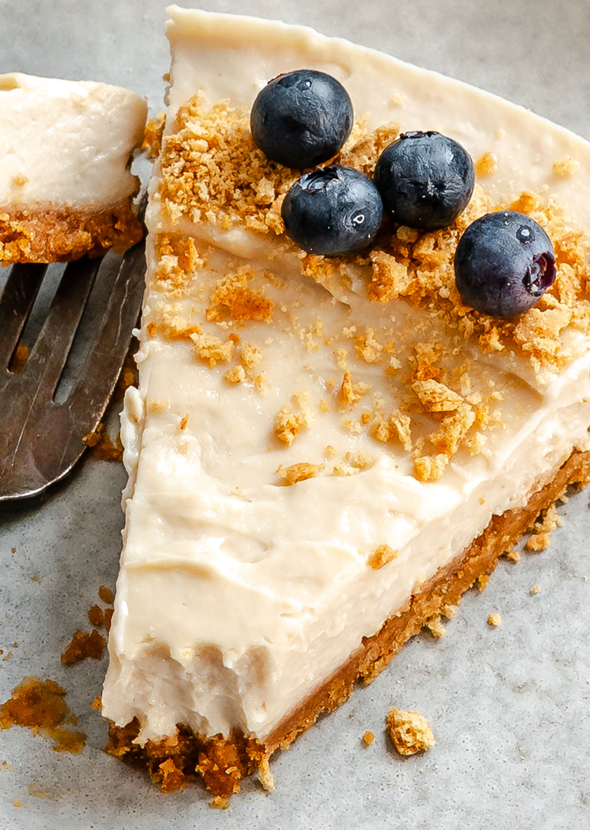 A slice of cheesecake with a crumbly crust, topped with blueberries and crumbled cookie pieces on a gray surface.