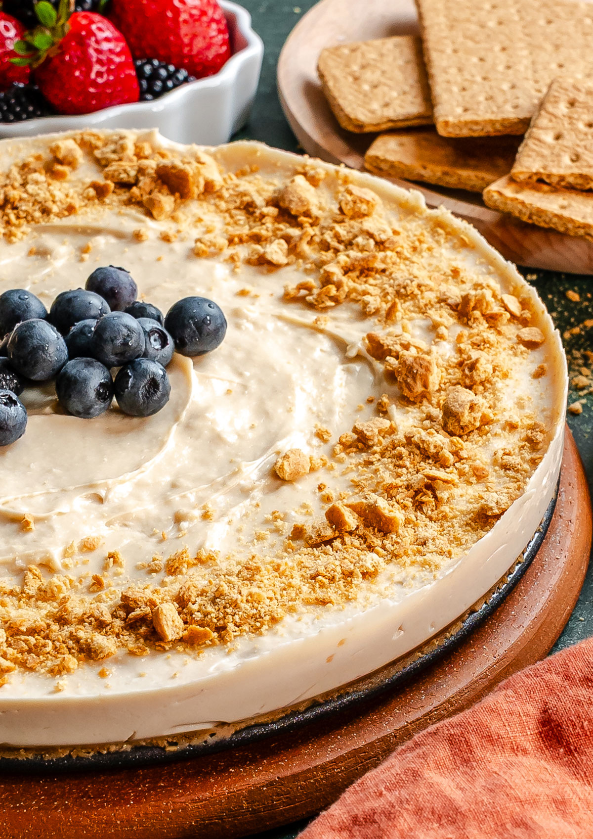 A cheesecake topped with crumbled graham crackers and blueberries, with a side of graham crackers and a bowl of assorted berries in the background.
