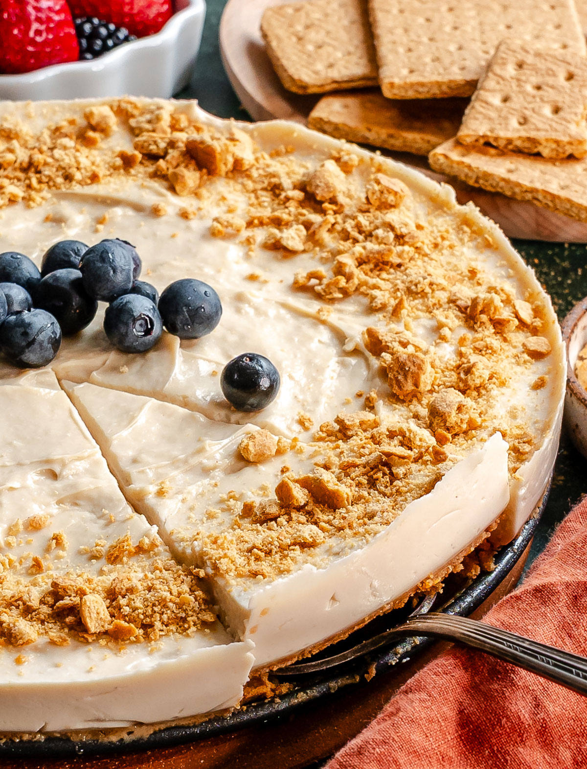 A round vegan cheesecake topped with blueberries and crumbled graham crackers, with one slice removed, next to graham crackers and a bowl of mixed berries.