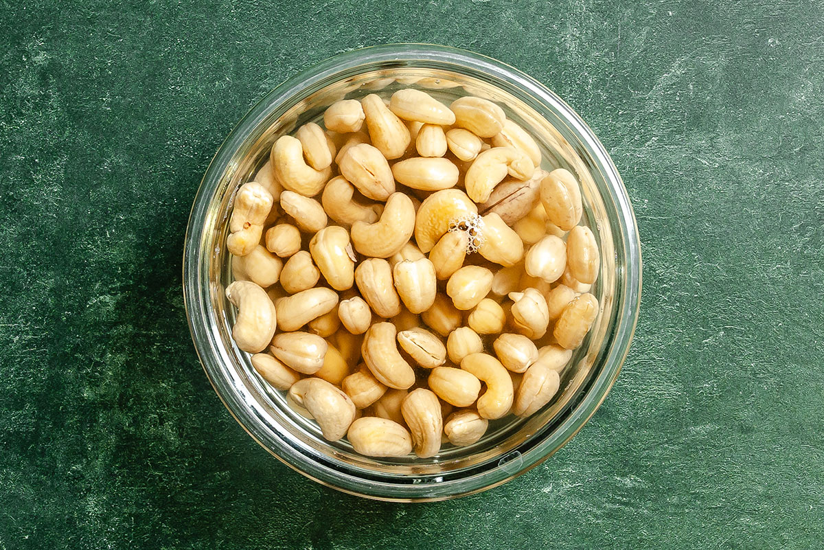 A glass bowl filled with soaked cashew nuts is placed on a green textured surface.