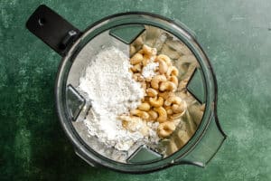 Overhead view of a blender containing cashews, flour, and a liquid ingredient, placed on a green surface.