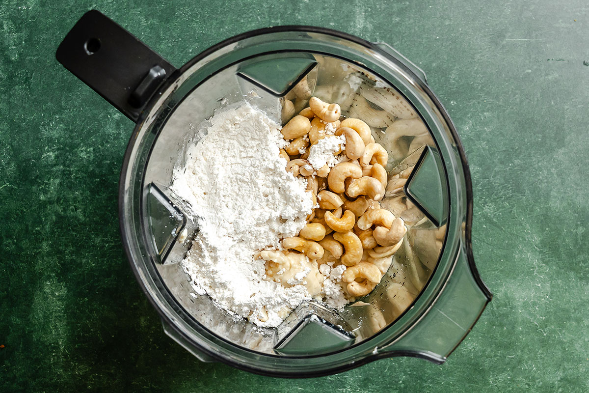 Overhead view of a blender containing cashews, flour, and a liquid ingredient, placed on a green surface.