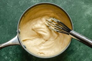 A saucepan filled with thick, creamy custard being stirred with a metal whisk on a green countertop.