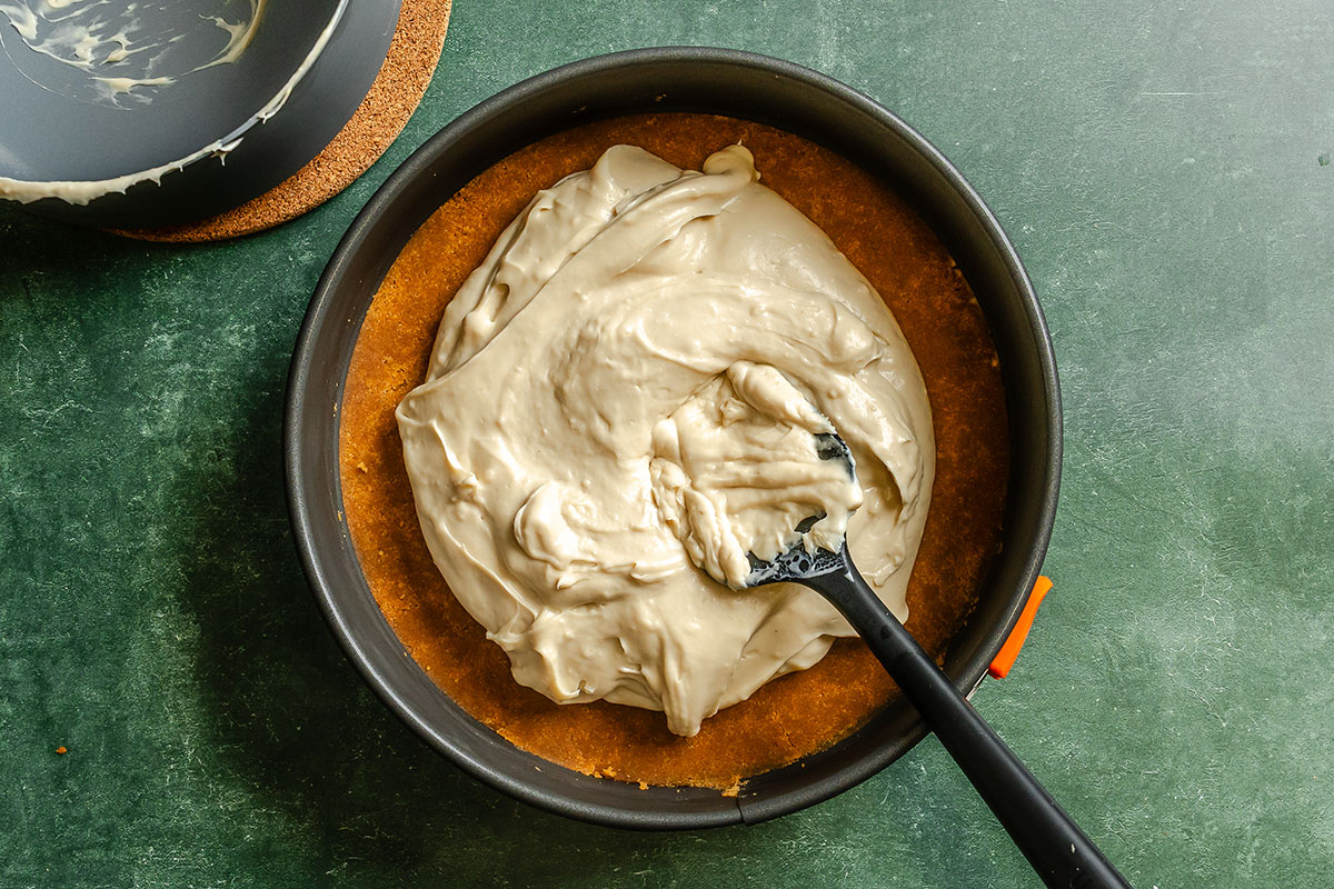 A spatula spreads cheesecake filling over a graham cracker crust in a round baking pan on a dark green surface.