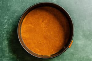 A round baking pan filled with an even layer of pressed graham cracker crust, viewed from above on a dark green surface.