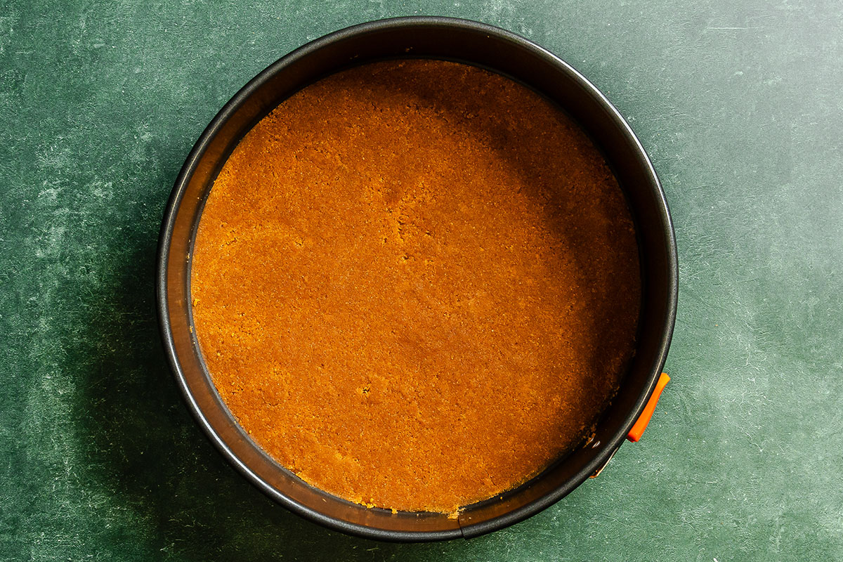 A round baking pan filled with an even layer of pressed graham cracker crust, viewed from above on a dark green surface.