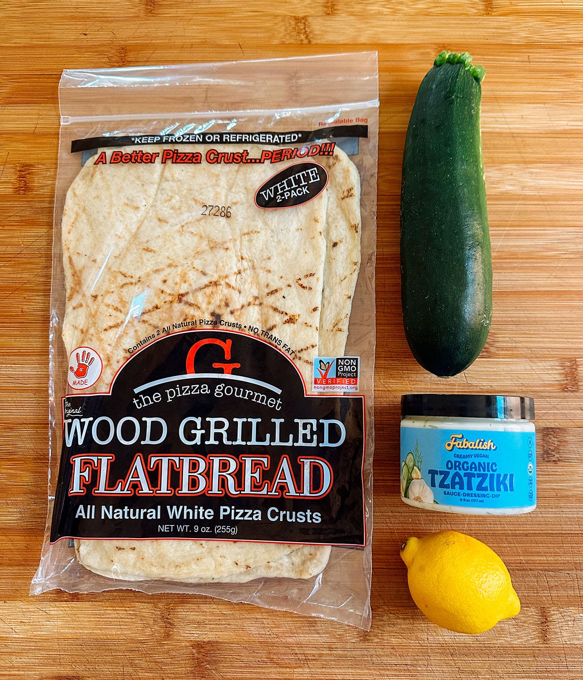 Packaged flatbread, a whole zucchini, a lemon, and a container of organic tzatziki are arranged on a wooden surface.