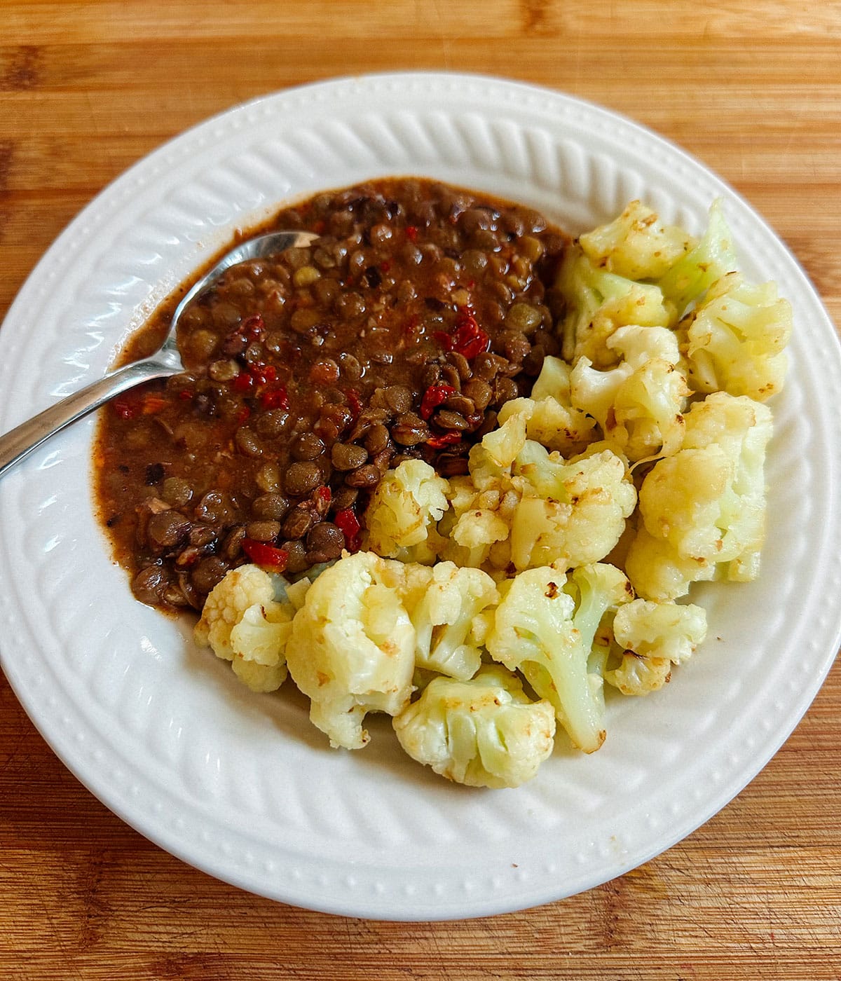 A white plate with cooked lentils in sauce and steamed cauliflower, placed on a wooden surface. A metal spoon rests in the lentils.