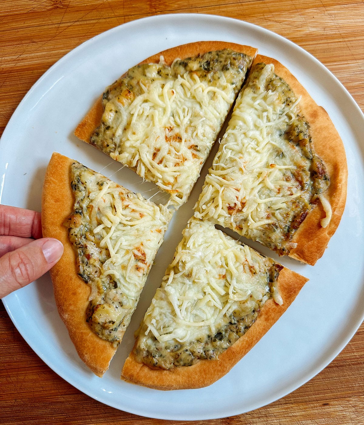 A hand holds a slice of flatbread topped with melted cheese and pesto, cut into four pieces and served on a white plate.