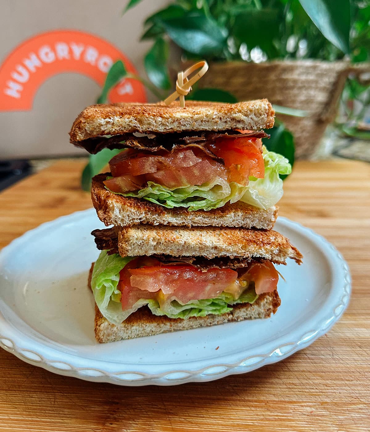 A stacked sandwich with lettuce, tomato, and bacon on toasted bread is served on a white plate, with a toothpick holding it together. A leafy plant and a Hungryroot box are in the background.