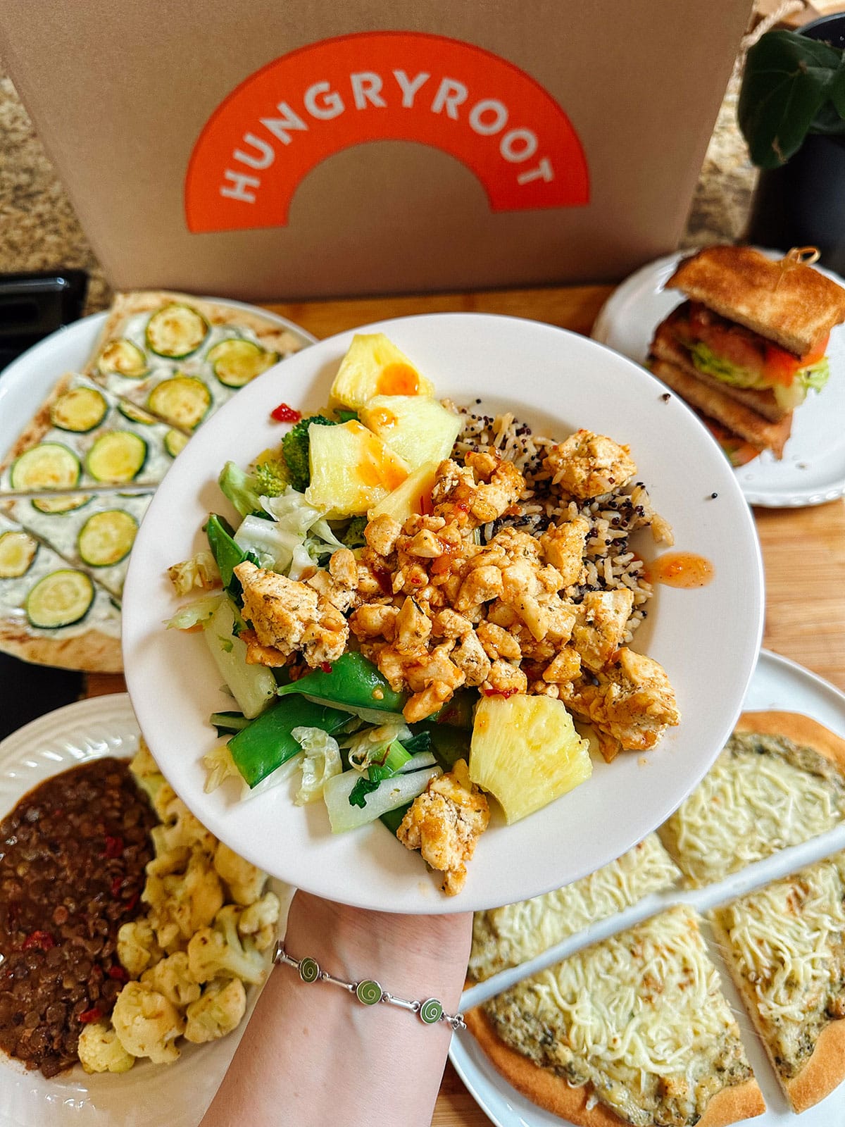 A person holds a bowl of mixed vegetables, pineapple, and crumbled protein in front of a Hungryroot box, with various prepared food dishes on a table.