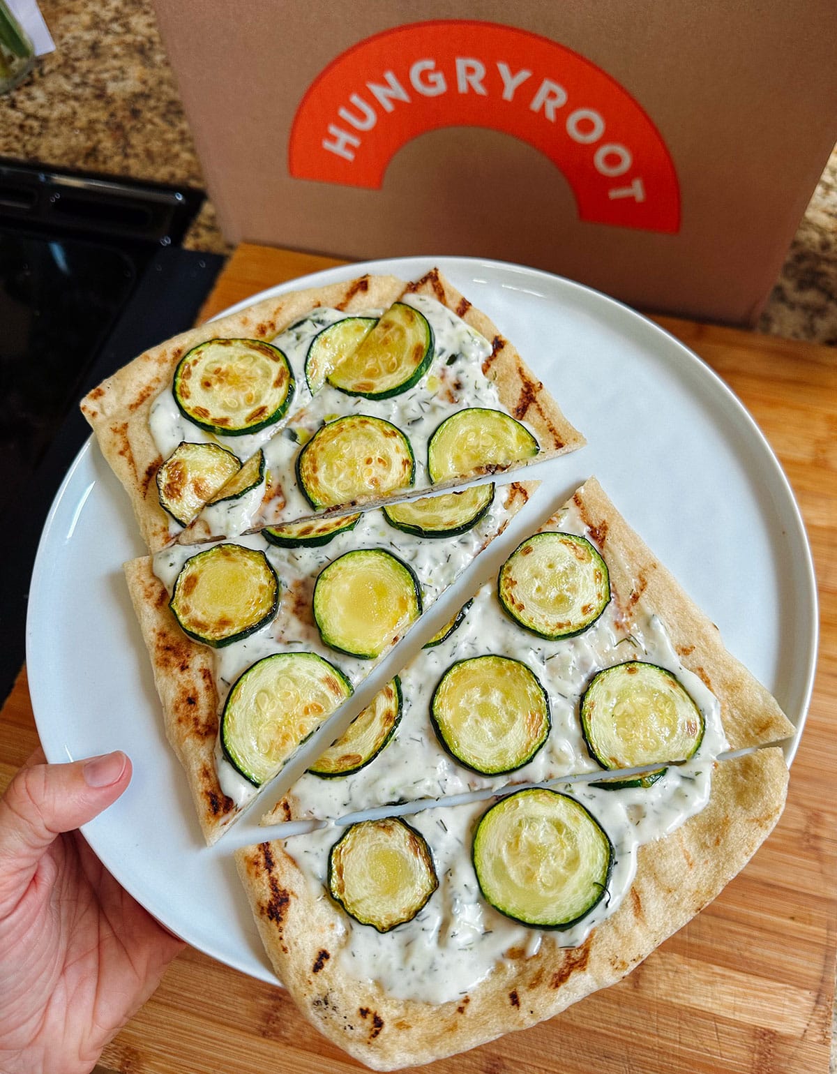 A hand holds a plate with a flatbread topped with sliced zucchini and white sauce, cut into sections. A Hungryroot box is in the background.