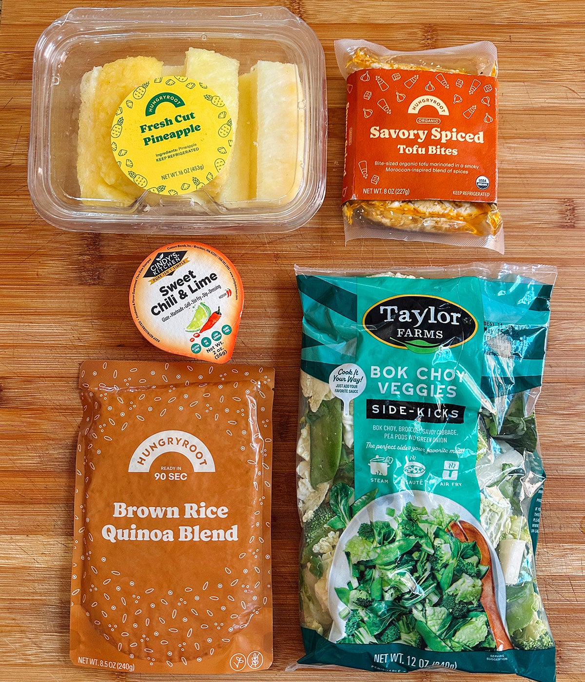 Five packaged food items on a wooden surface: fresh cut pineapple, savory spiced tofu bites, sweet chili lime sauce, brown rice quinoa blend, and a bag of bok choy veggies.