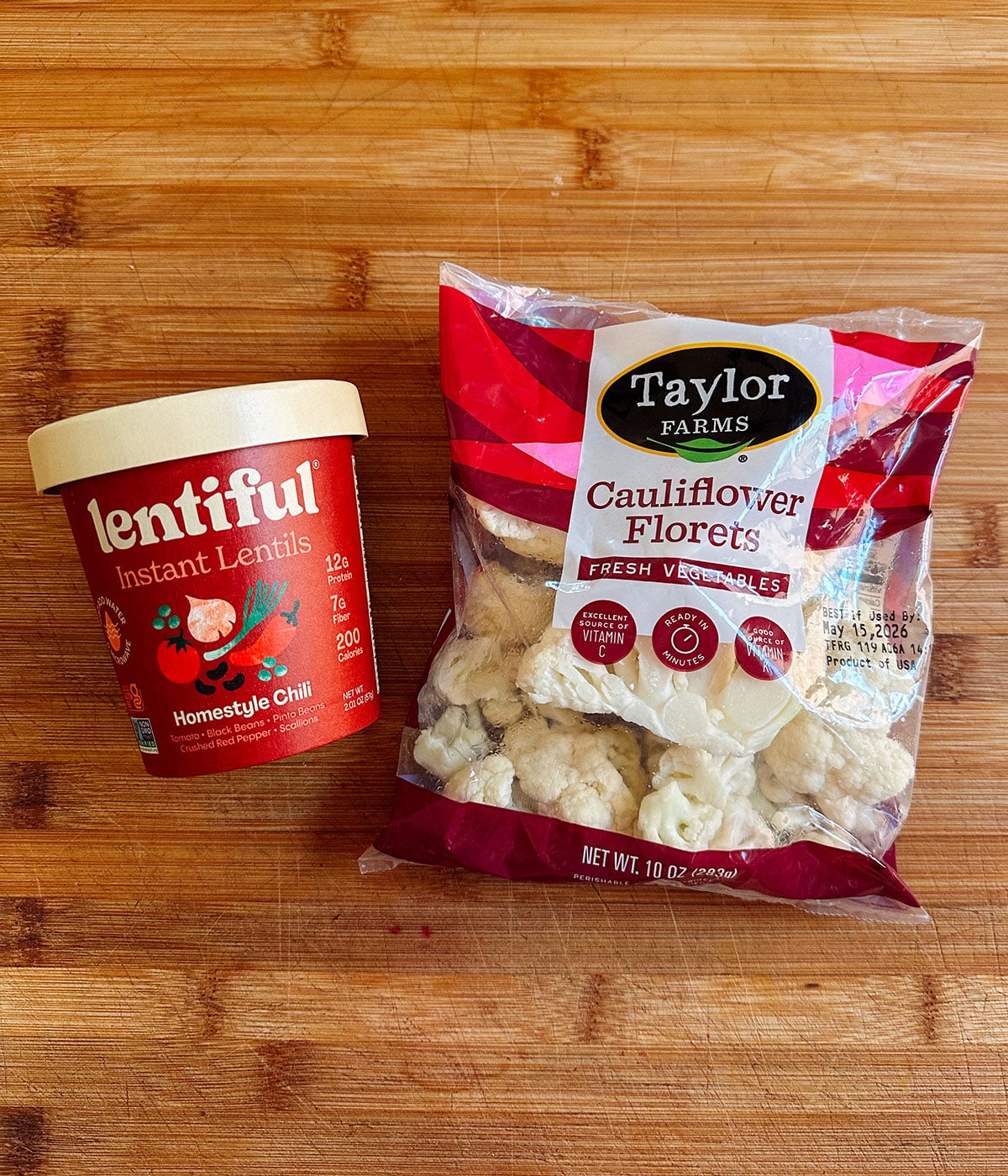 A container of Lentiful Instant Lentils Homestyle Chili next to a bag of Taylor Farms Cauliflower Florets on a wooden surface.
