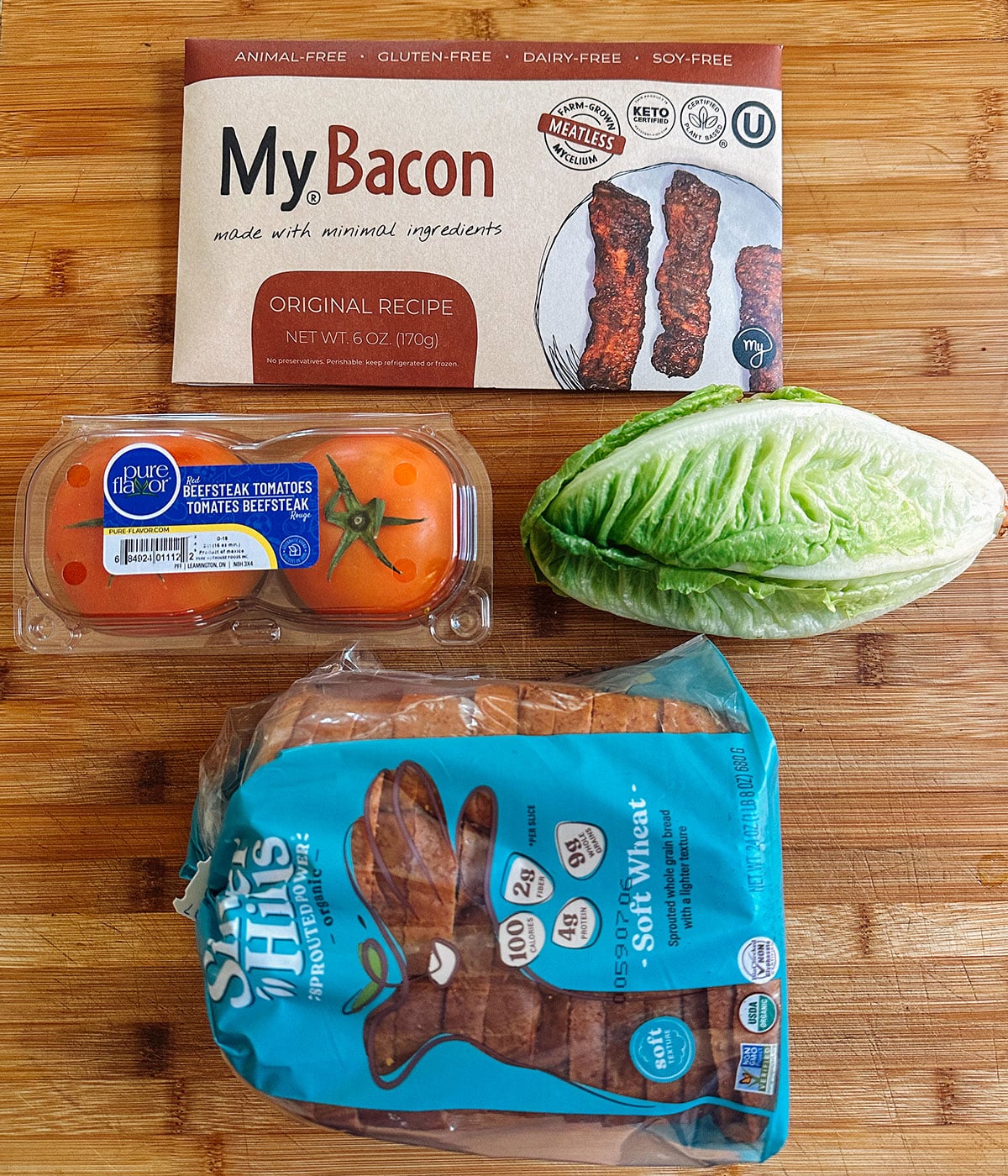 A package of plant-based bacon, grape tomatoes, a romaine lettuce heart, and a bag of sliced wheat sandwich buns on a wooden surface.