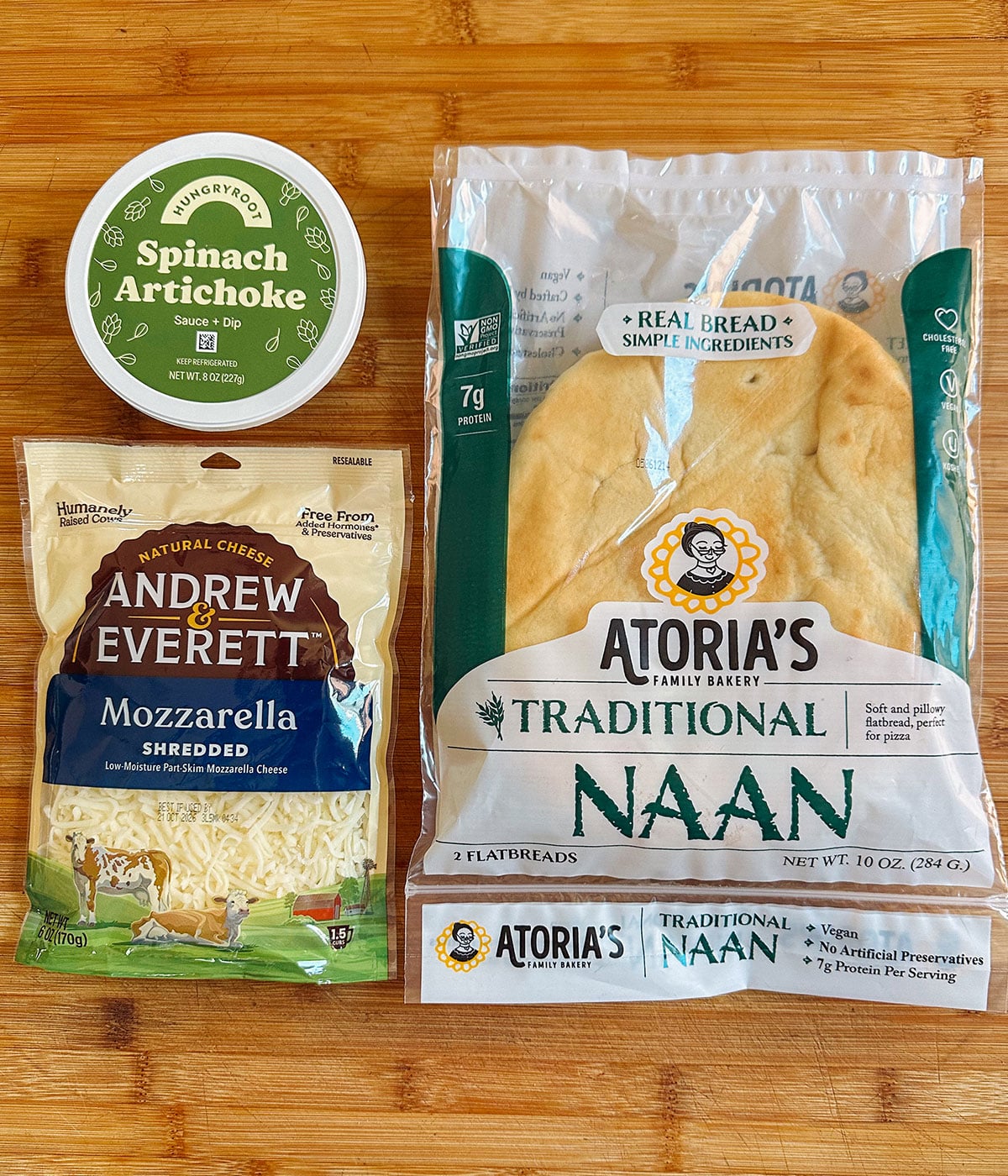 A container of spinach artichoke dip, a package of shredded mozzarella cheese, and a package of traditional naan on a wooden surface.