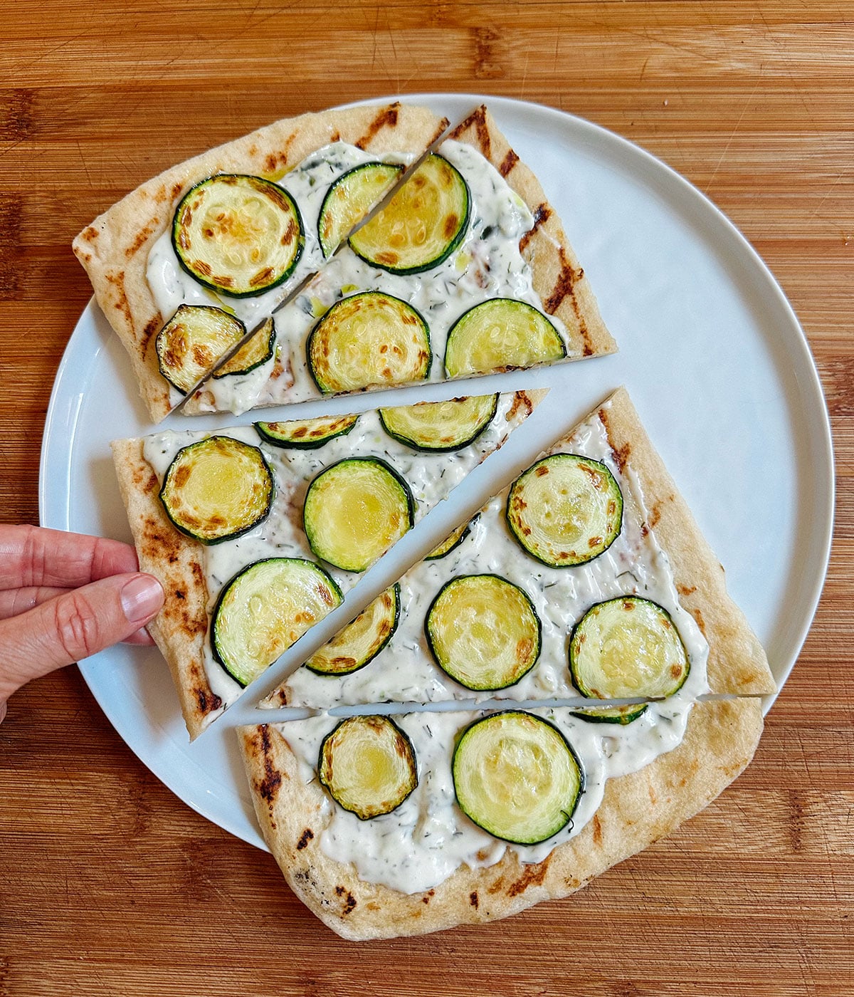 A white plate with flatbread topped with white sauce and zucchini slices, cut into three triangles, on a wooden surface. A hand is holding one slice.