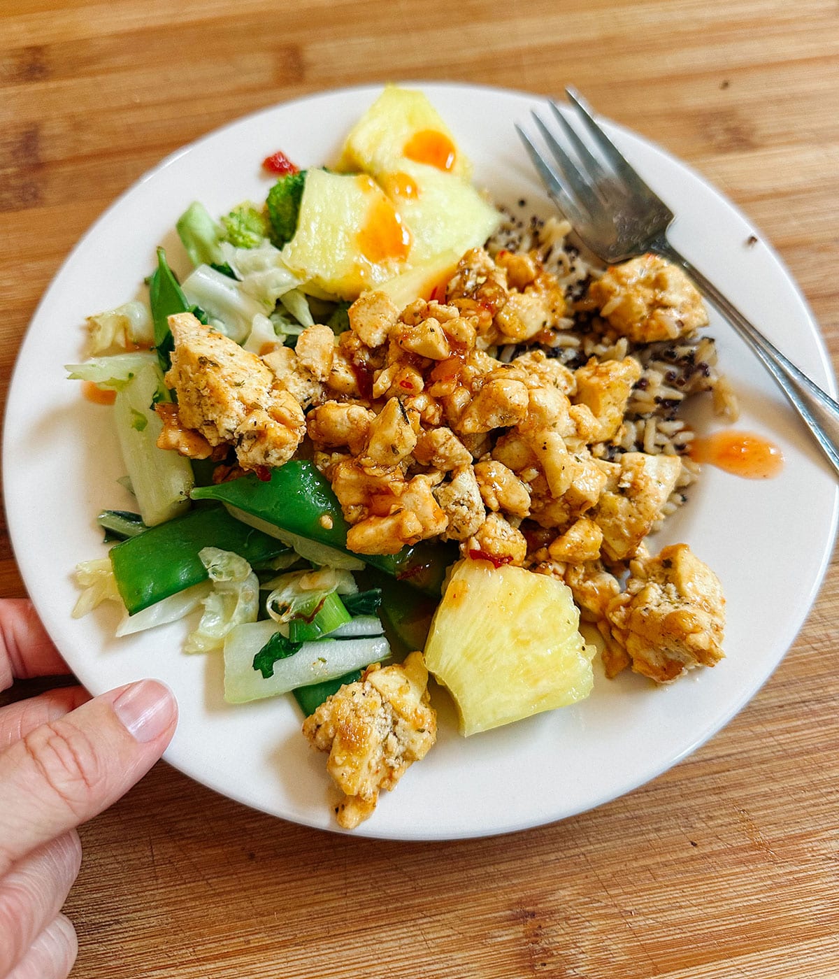 A white plate with cooked chicken, rice, steamed vegetables, pineapple chunks, and a drizzle of orange sauce, on a wooden surface with a fork and a hand holding the plate.
