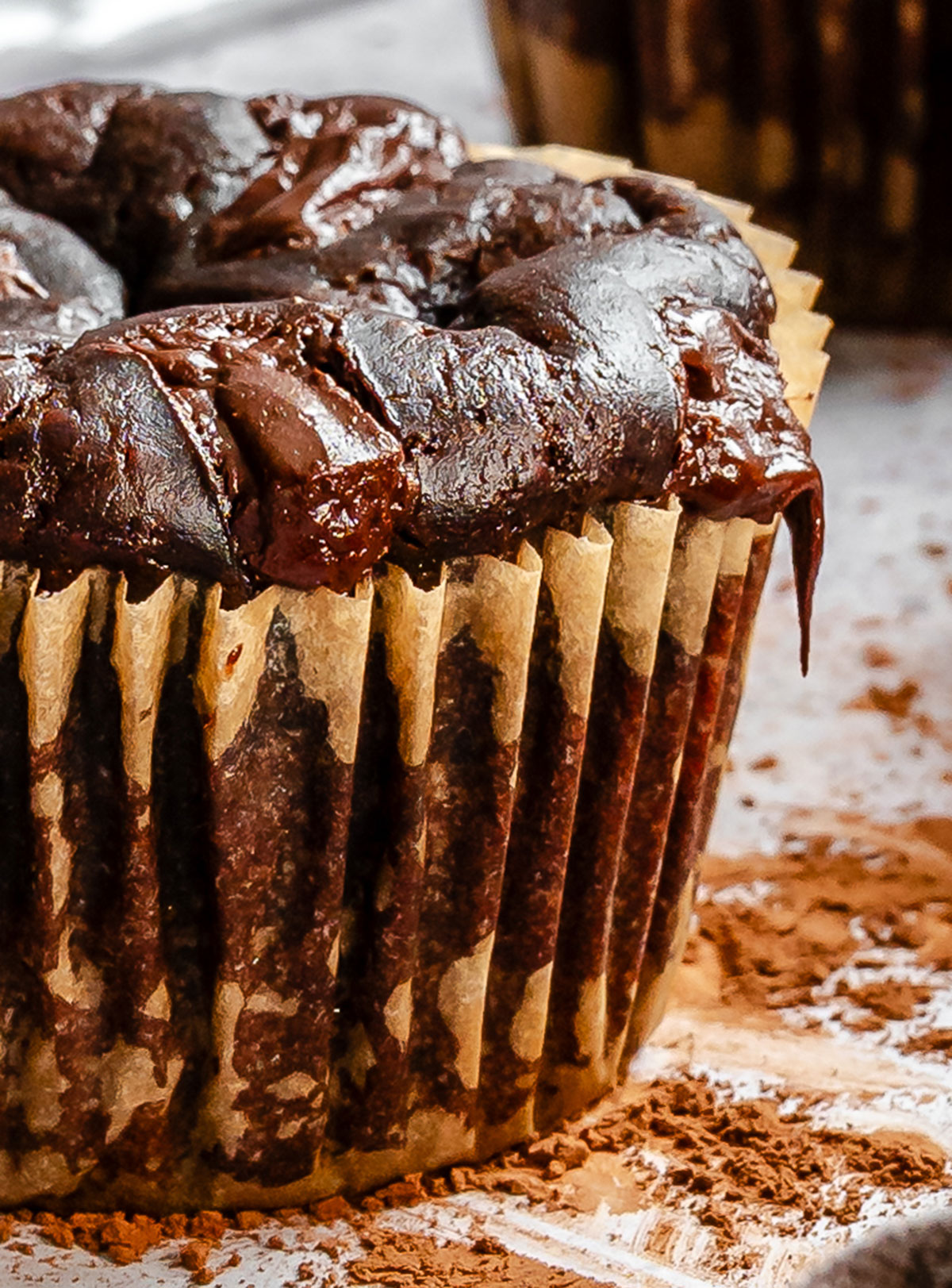 Close-up of a chocolate muffin with a swirl of rich, glossy chocolate topping in a brown paper wrapper, dusted cocoa powder visible on the surface nearby.