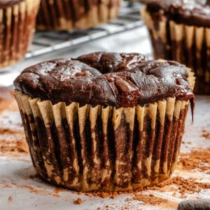 A close-up of a chocolate muffin in a paper liner, with more muffins and cocoa powder in the background.