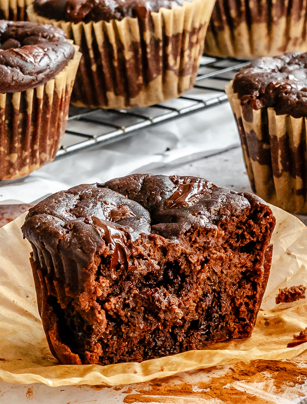 A close-up of a chocolate muffin with a bite taken out, showing a moist, dense interior. Other whole muffins in paper liners are in the background on a cooling rack.