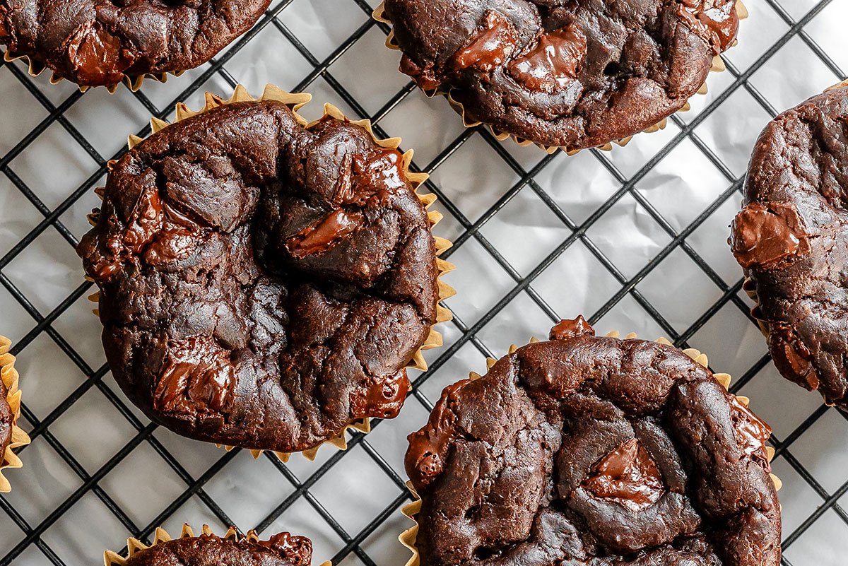 Chocolate muffins with chunks of chocolate rest on a black wire cooling rack lined with parchment paper.
