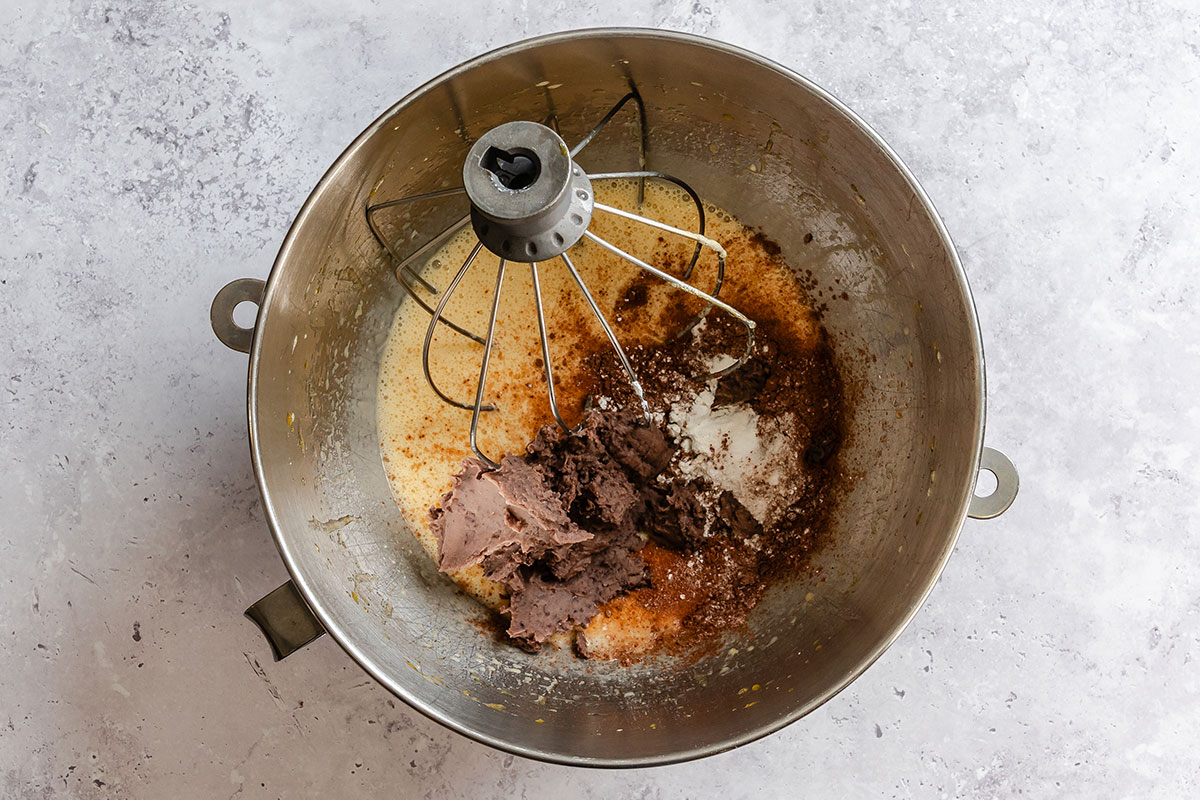Overhead view of a metal mixing bowl with a whisk attachment containing partially mixed wet and dry baking ingredients, including cocoa powder and creamed butter.