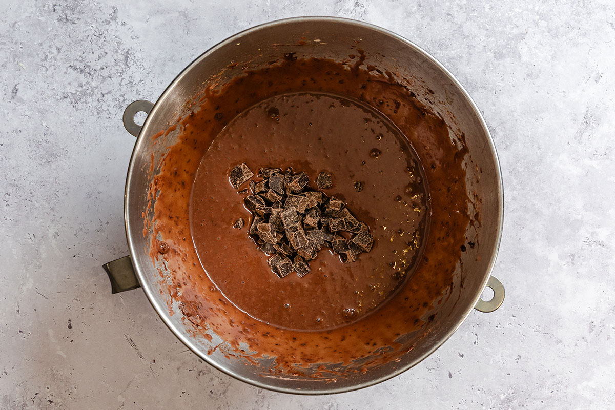 A metal mixing bowl containing chocolate cake batter with chunks of chocolate on top, placed on a light gray surface.
