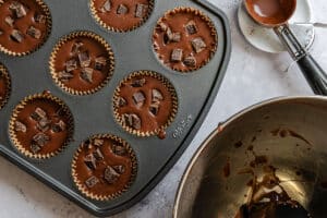 A muffin tray filled with chocolate batter and chocolate chunks, a metal mixing bowl with residue, and a metal measuring cup with chocolate nearby.
