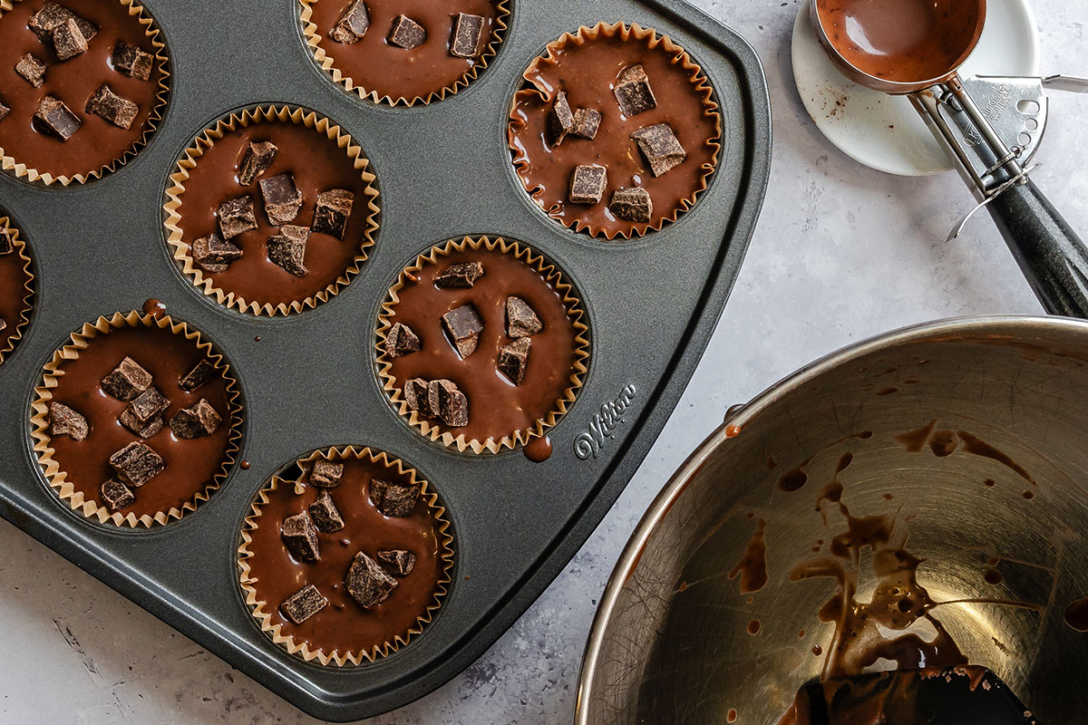 A muffin tray filled with chocolate batter and chocolate chunks, a metal mixing bowl with residue, and a metal measuring cup with chocolate nearby.