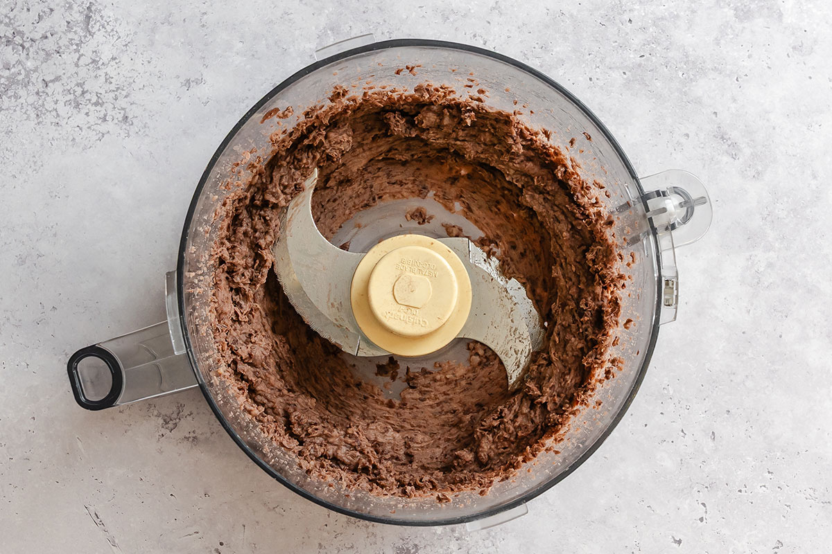 A food processor containing a mixture of blended black beans, viewed from above on a light-colored surface.