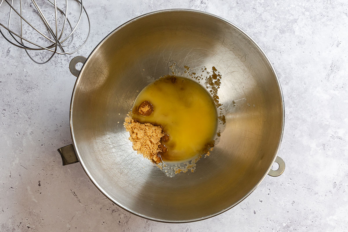A metal mixing bowl with melted butter and brown sugar, next to a metal whisk, on a light grey countertop.