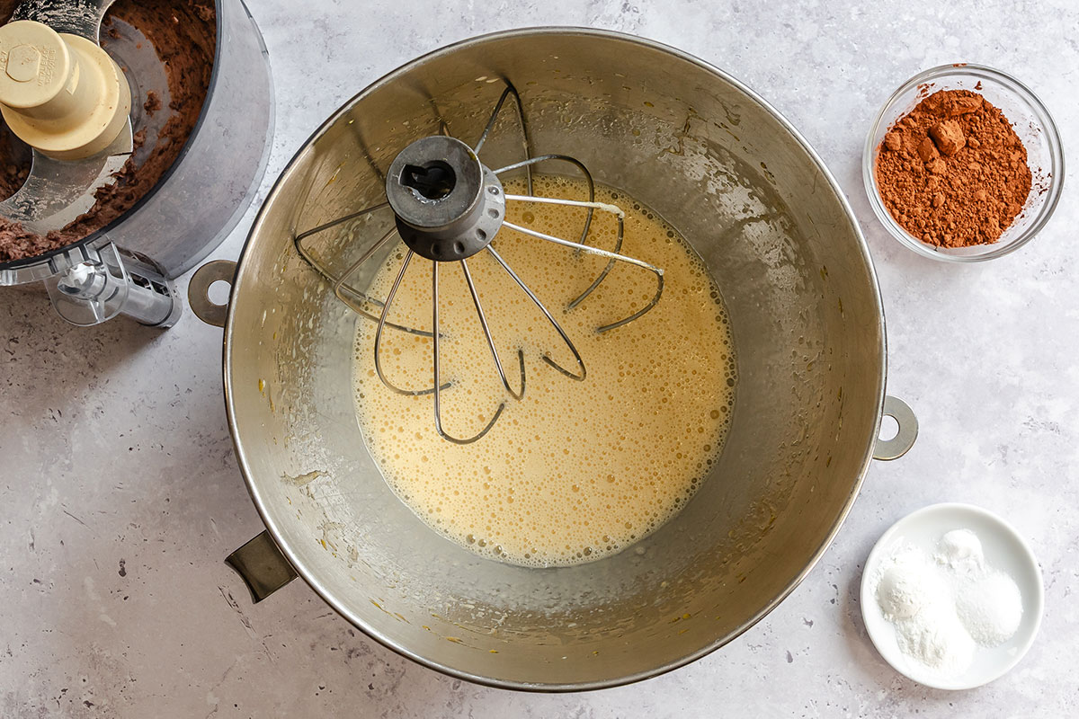 A mixing bowl with a whisk attachment contains a beaten egg mixture. Nearby are a food processor with chocolate batter, a bowl of cocoa powder, and a small dish with baking soda and powder.