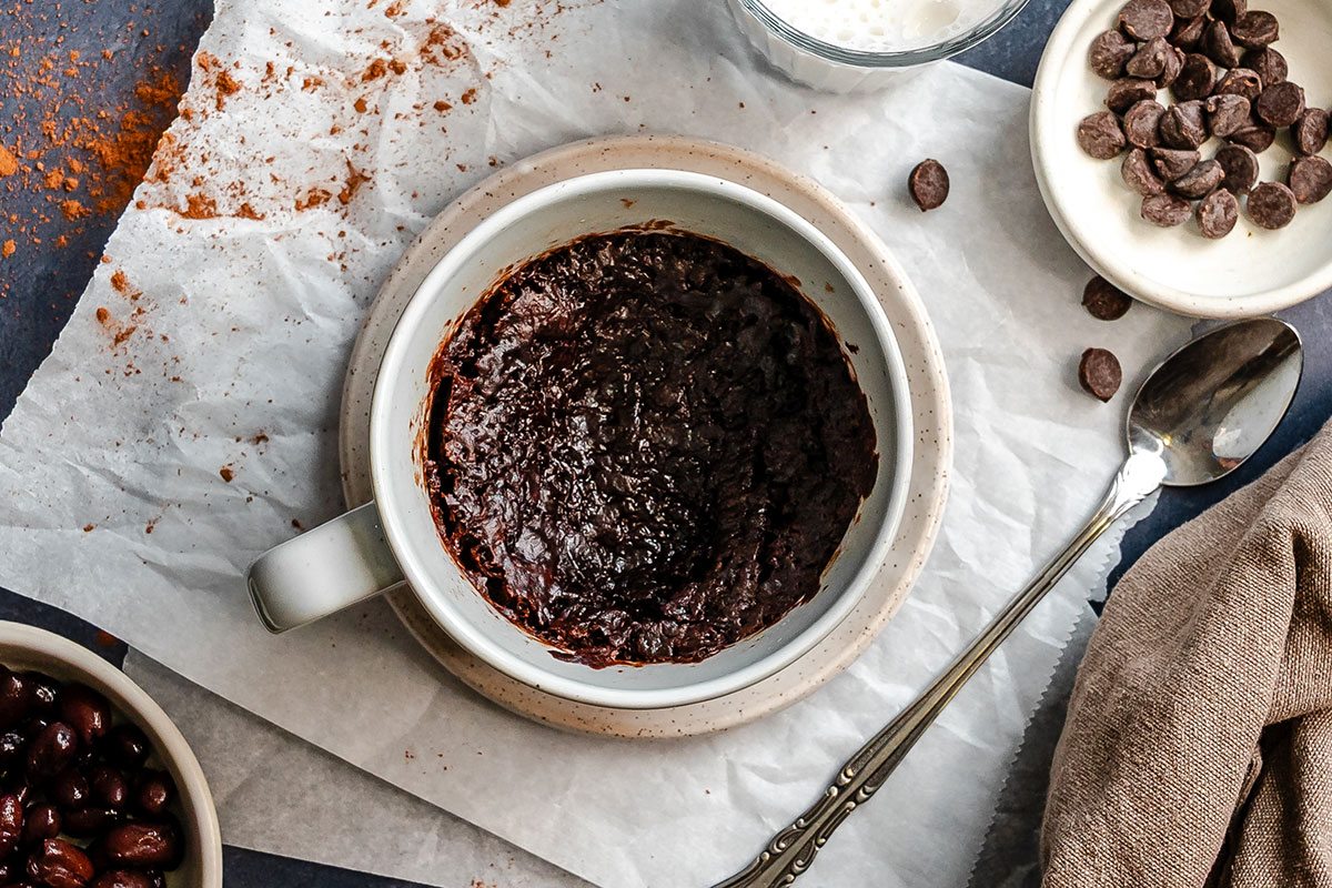 A mug filled with baked chocolate cake sits on parchment paper, surrounded by chocolate chips, a glass of milk, and a spoon.