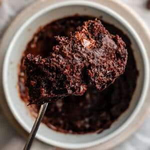 A close-up of a spoon holding a portion of chocolate mug cake above a white ramekin filled with the same dessert.